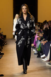 Model walks the runway in a black floral-embroidered coat with a belt, paired with black boots.
