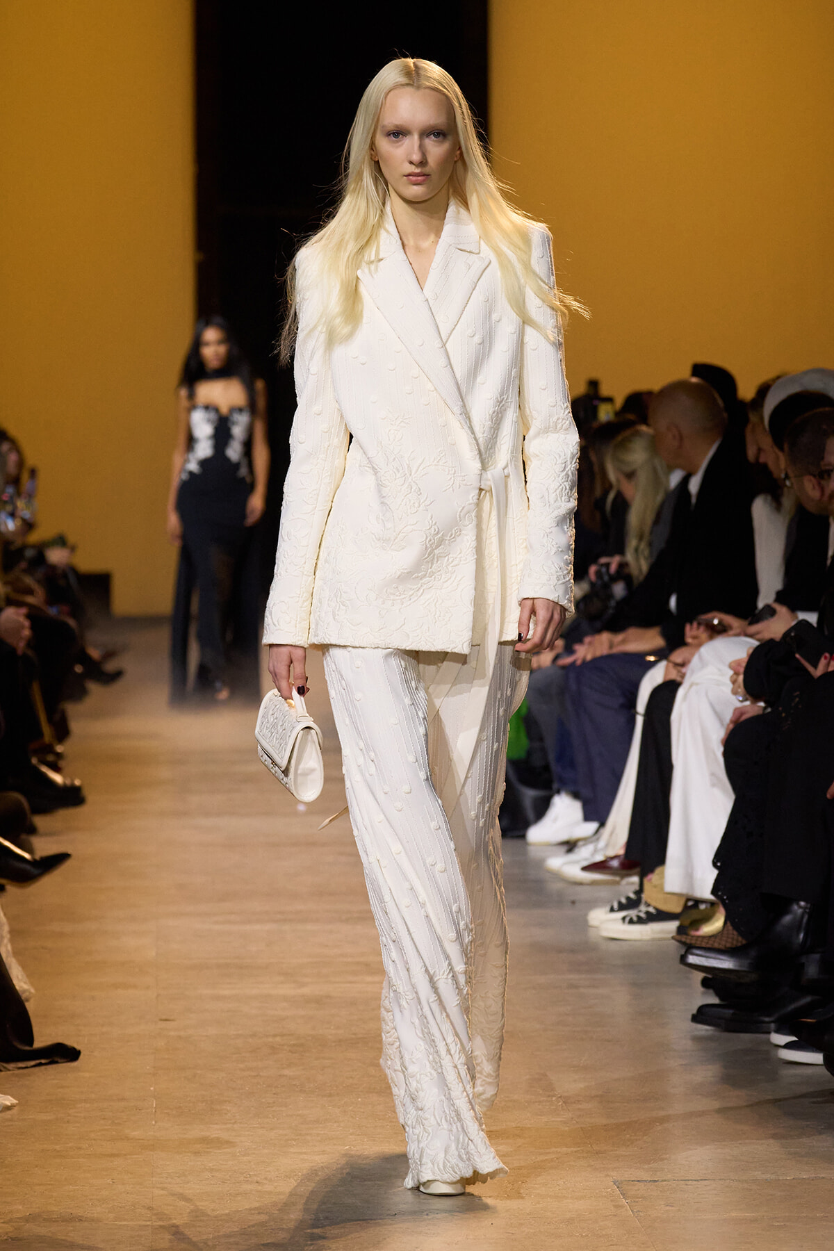 Slim model walking a fashion runway in an all‑white textured suit, holding a small white clutch.