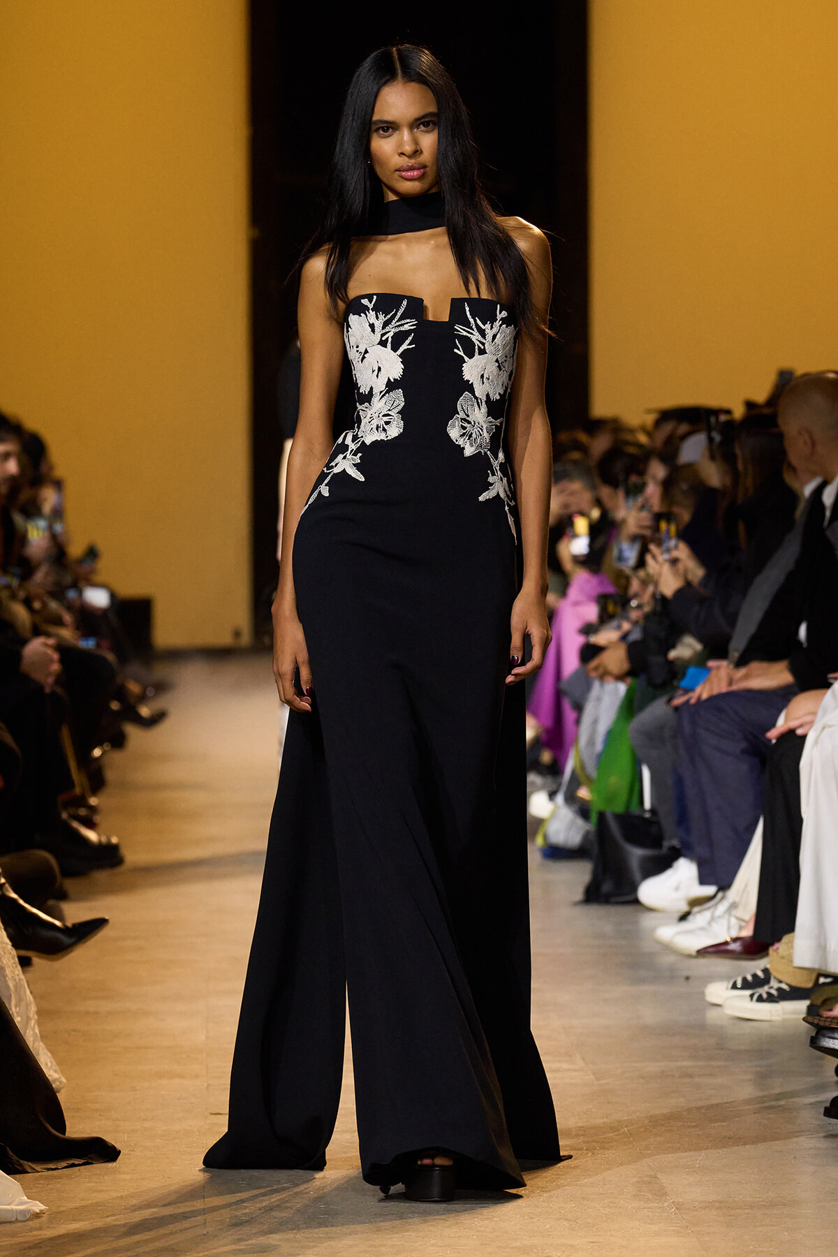 Model walks a fashion runway in a black gown with white floral embroidery along the bodice, audience seated and watching on both sides.