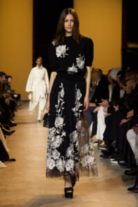 Model walks the runway in a black knit top with large white floral embroidery and a sheer black floral-embroidered skirt, holding a small black handbag; audience seated along the catwalk in the background.