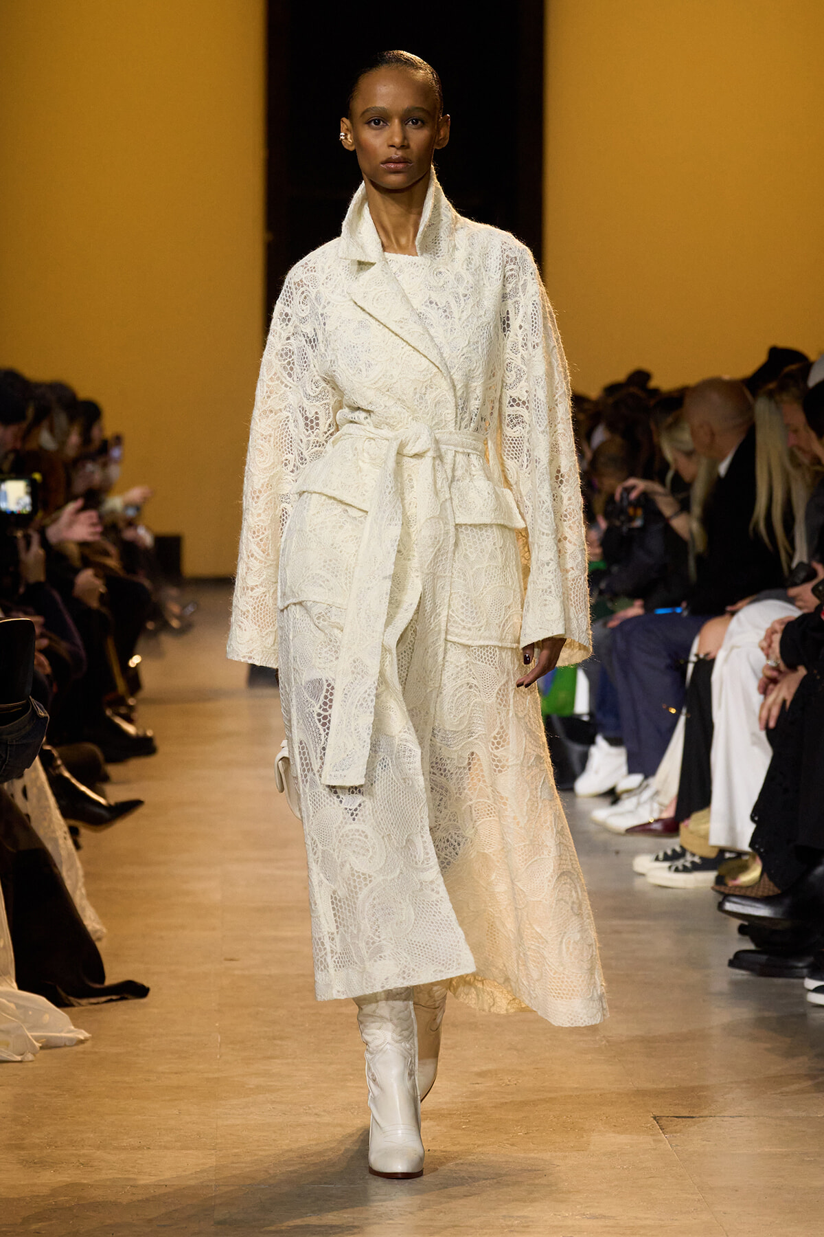 Model walks a fashion runway wearing a long white lace coat with a tied belt and matching white boots, audience visible on both sides of the catwalk.
