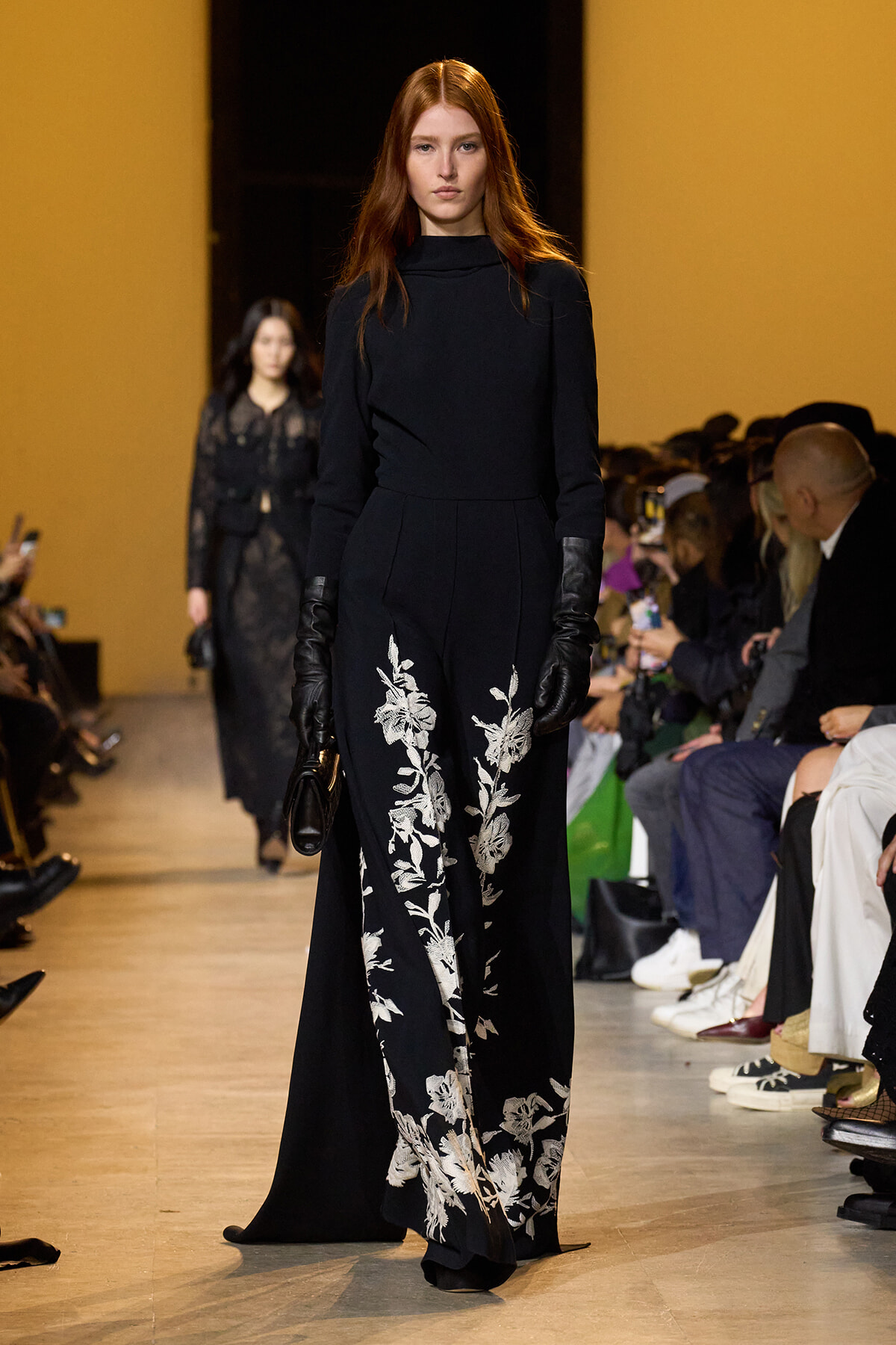 Model walking the runway in a black high-neck gown with silver floral embroidery, paired with black gloves. Audience visible on the sides.] , but let's ensure it's clean.