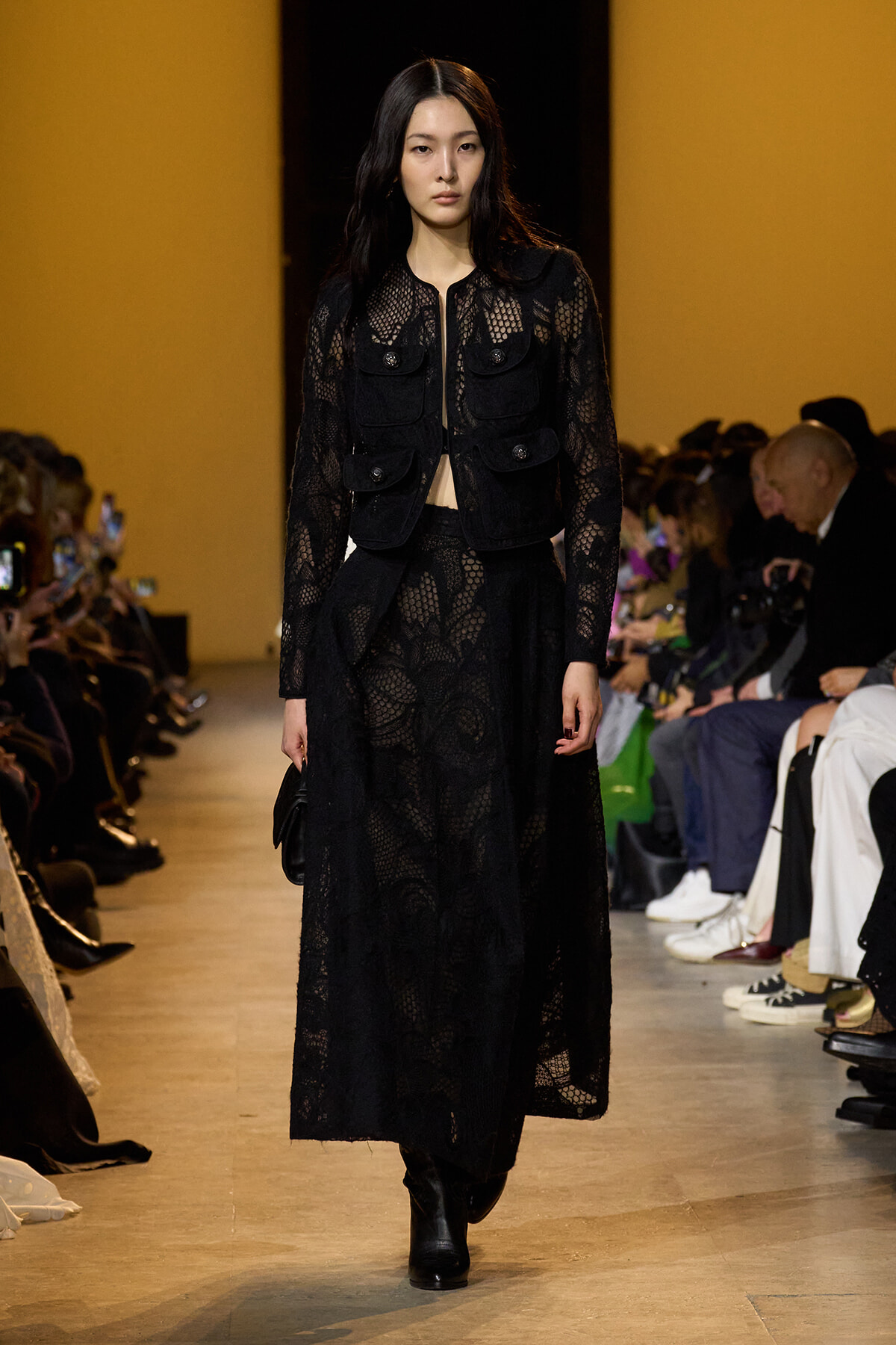 Model walks a fashion runway in a black lace ensemble, with a sheer long dress and cropped jacket, audience on both sides.