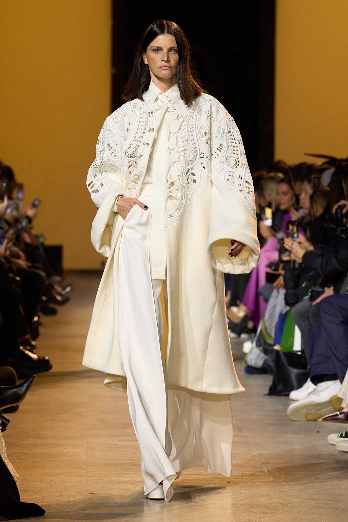 Model on a runway wearing a white, lacy cape over a cream suit; audience seated along the catwalk.