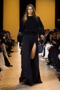 Model walking a fashion runway in a black lace-sleeved sweater and a long black skirt with a high slit; audience lines the catwalk in a dimly lit venue.