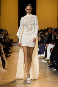 Model walks the runway in a white, long-sleeve lace-detailed dress with a flowing cape and high neckline, audience in background.