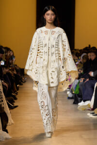 Model walks a fashion show wearing an intricate white lace cape and matching pants with cut-out patterns; audience seated along the runway.