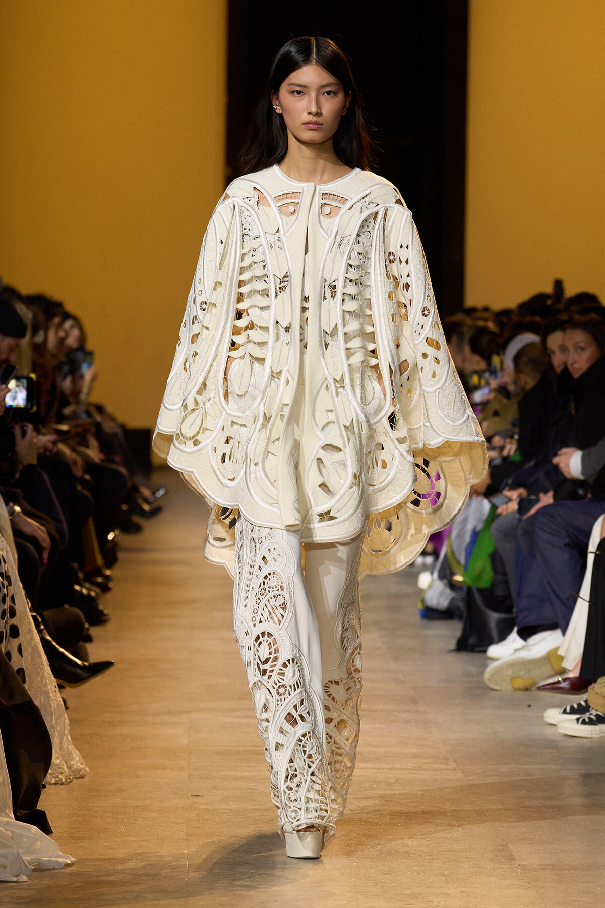 Model walks a fashion show wearing an intricate white lace cape and matching pants with cut-out patterns; audience seated along the runway.