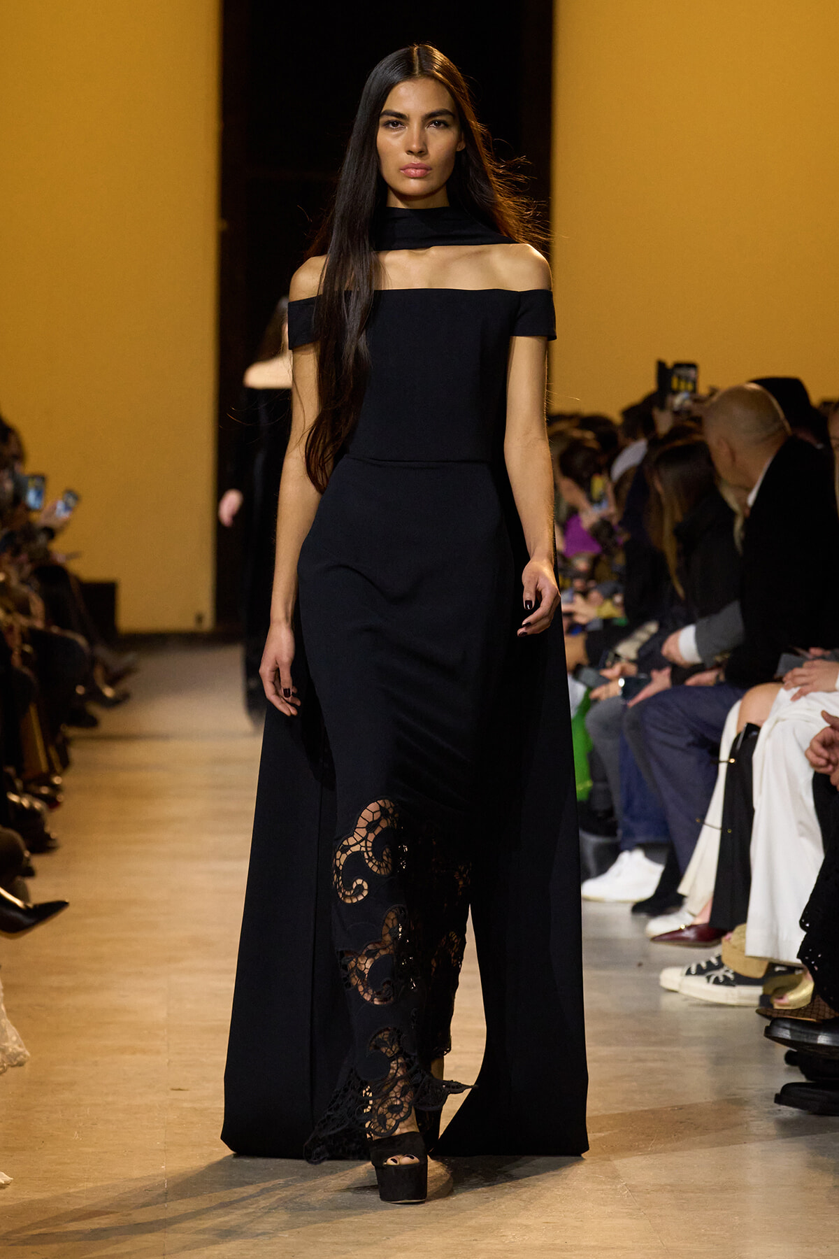 Model walking a runway in a floor-length black off-shoulder gown with lace panels; audience seated along the sides of the catwalk.