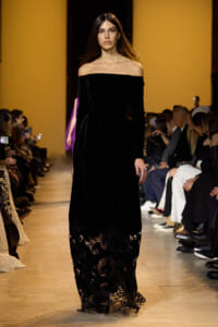 Model walks the runway in a long off-the-shoulder black velvet gown with lace trim, audience seated along both sides of the catwalk.