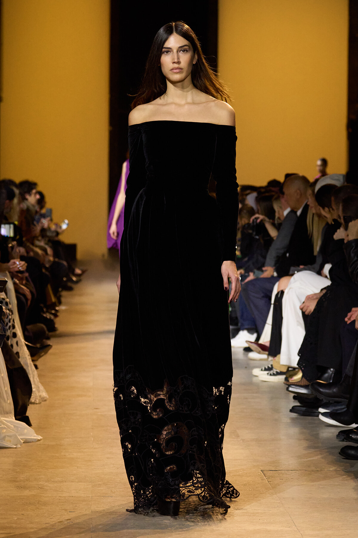 Model walks the runway in a long off-the-shoulder black velvet gown with lace trim, audience seated along both sides of the catwalk.