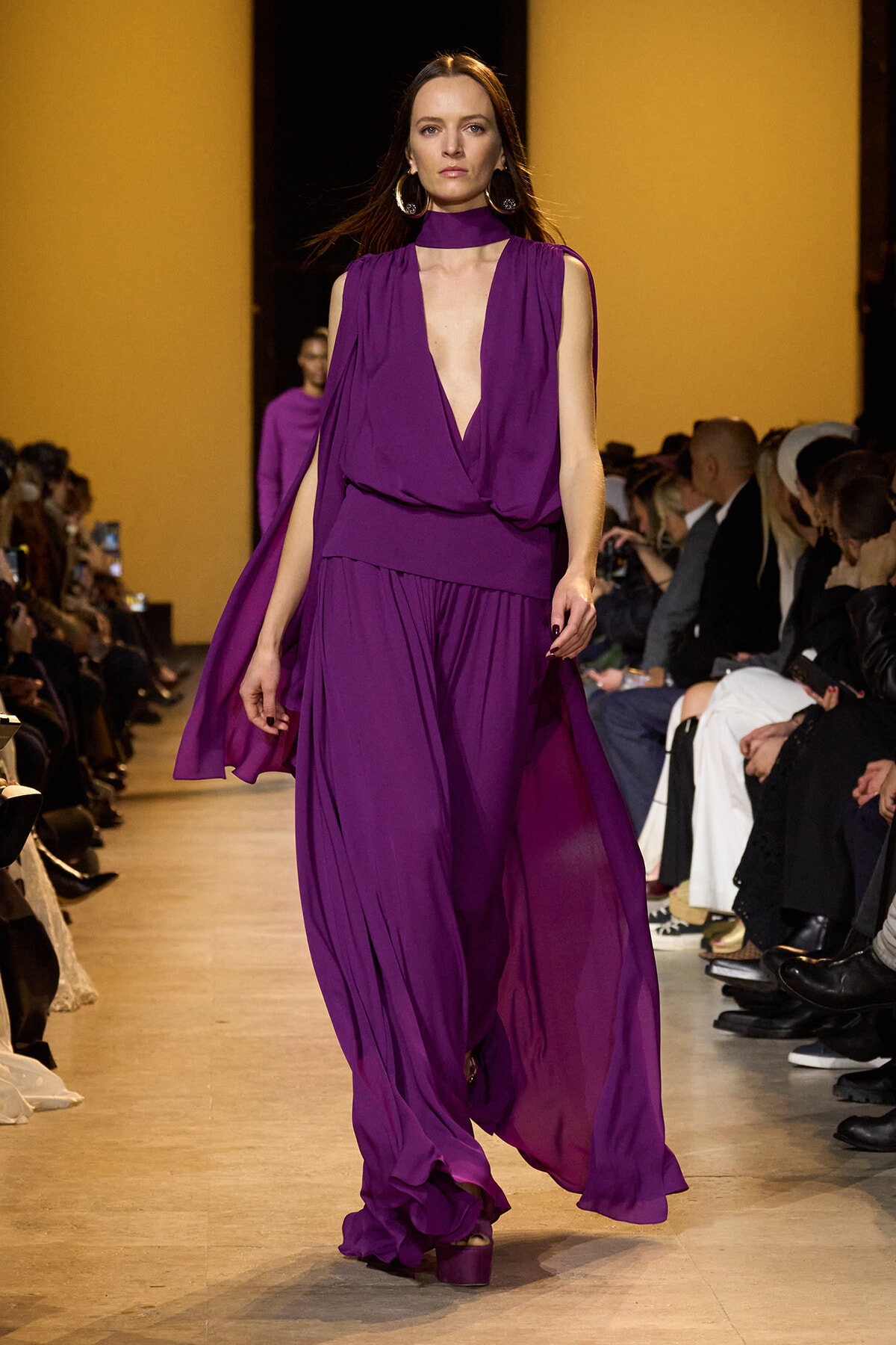 Model walks the runway in a voluminous purple gown with a deep V neckline and choker-style top, audience on both sides watching.