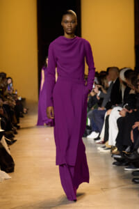 Model walking runway in a purple draped blouse and wide-leg pants; bold magenta ensemble with audience on both sides of the catwalk.