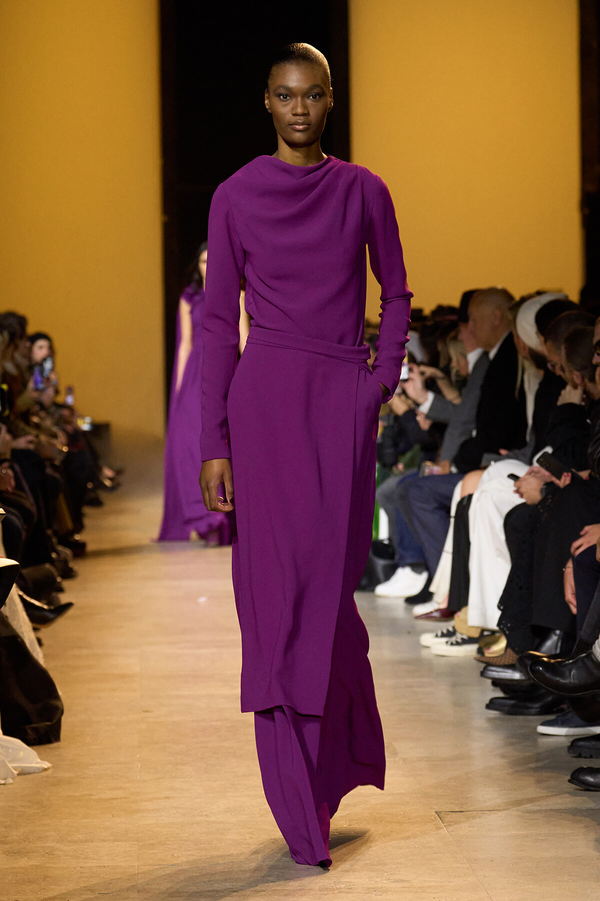 Model walking runway in a purple draped blouse and wide-leg pants; bold magenta ensemble with audience on both sides of the catwalk.