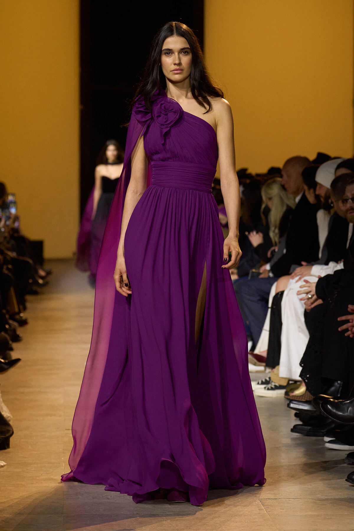 Model on runway wearing a one-shoulder purple gown with a large floral shoulder detail.