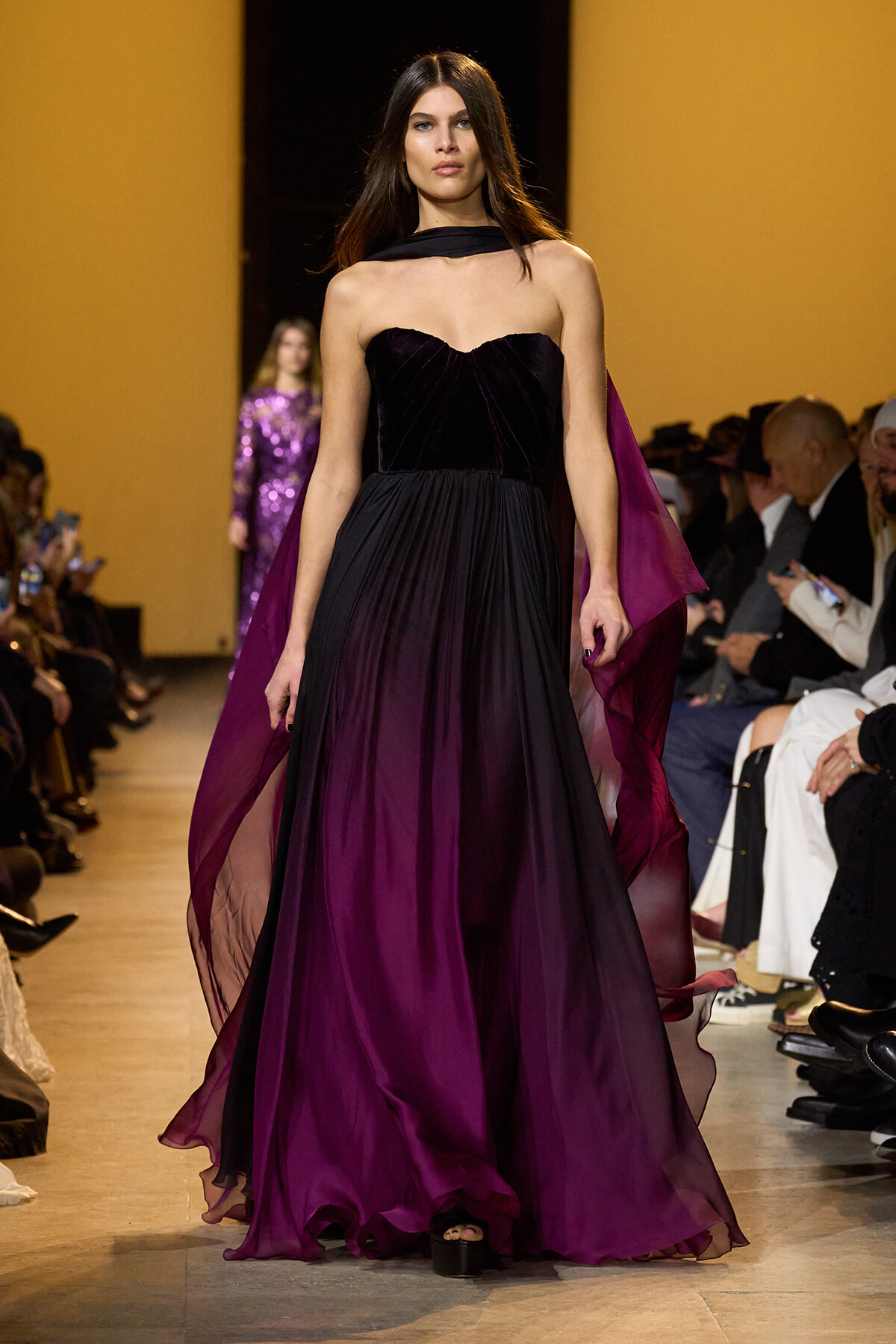 Model walking the runway in a floor-length, gradient purple gown with a black velvet bodice and sheer cape on a fashion show catwalk.
