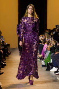 Model walks a runway in a long purple lace dress with floral patterns and matching platform heels, audience watching on both sides.