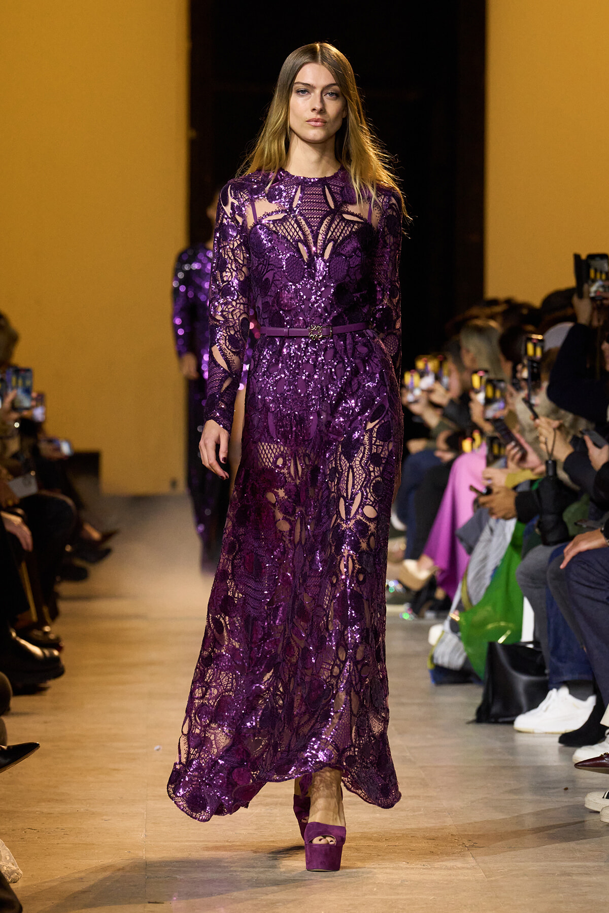 Model walks a runway in a long purple lace dress with floral patterns and matching platform heels, audience watching on both sides.