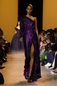 Model on a fashion runway wearing a one-shoulder, floor-length purple sequin gown with a thigh-high slit.