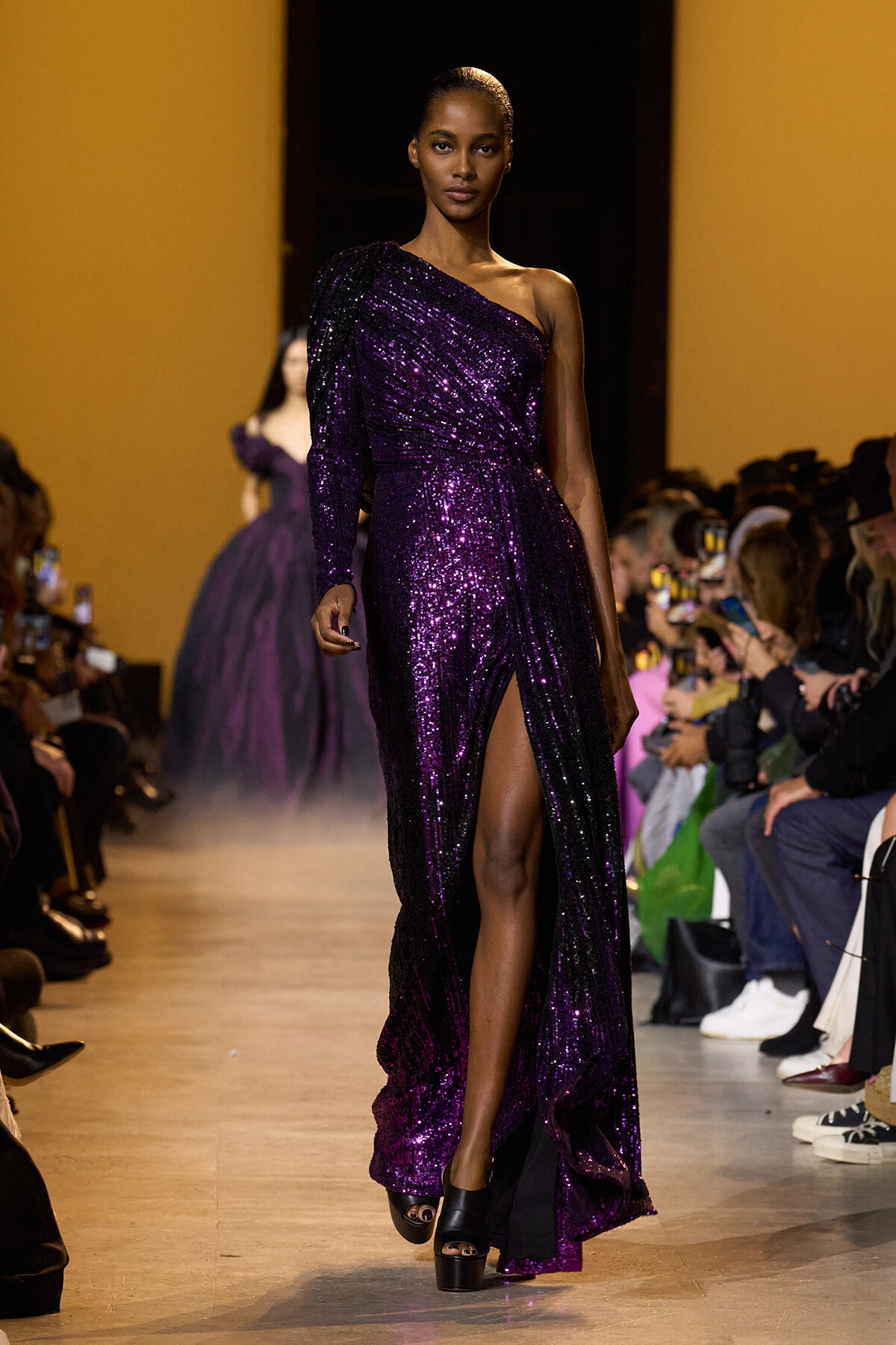 Model on a fashion runway wearing a one-shoulder, floor-length purple sequin gown with a thigh-high slit.