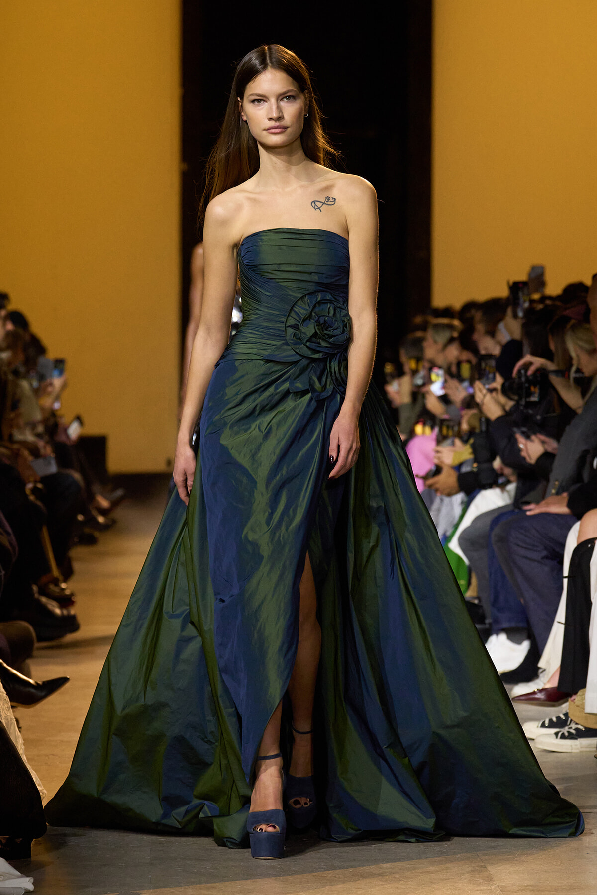Model walks the runway in a strapless deep teal gown with a large fabric flower, a long draped train, and blue platform heels; audience lines the sides.