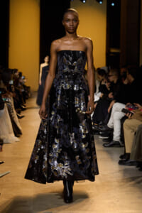 Model walks the runway in a strapless black gown with silver-blue sequined floral patterns, audience blurred in the background.
