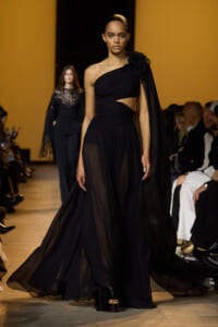 Model walks the runway in a black one-shoulder gown with a floral shoulder detail and flowing sheer skirt.