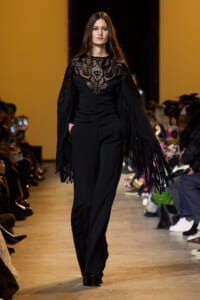 Model on a runway wearing a black lace-embellished top with long fringe sleeves and matching black trousers; audience watches from sides.