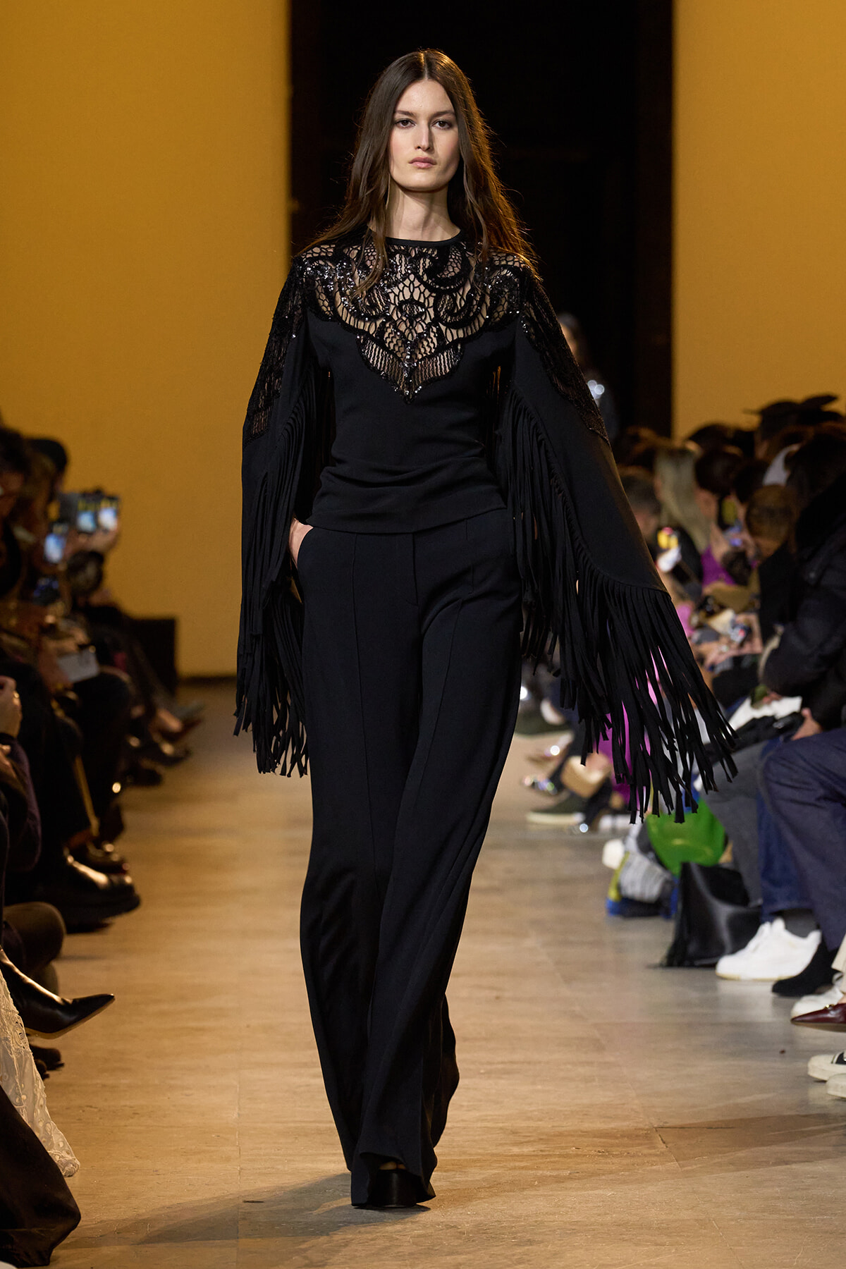 Model on a runway wearing a black lace-embellished top with long fringe sleeves and matching black trousers; audience watches from sides.