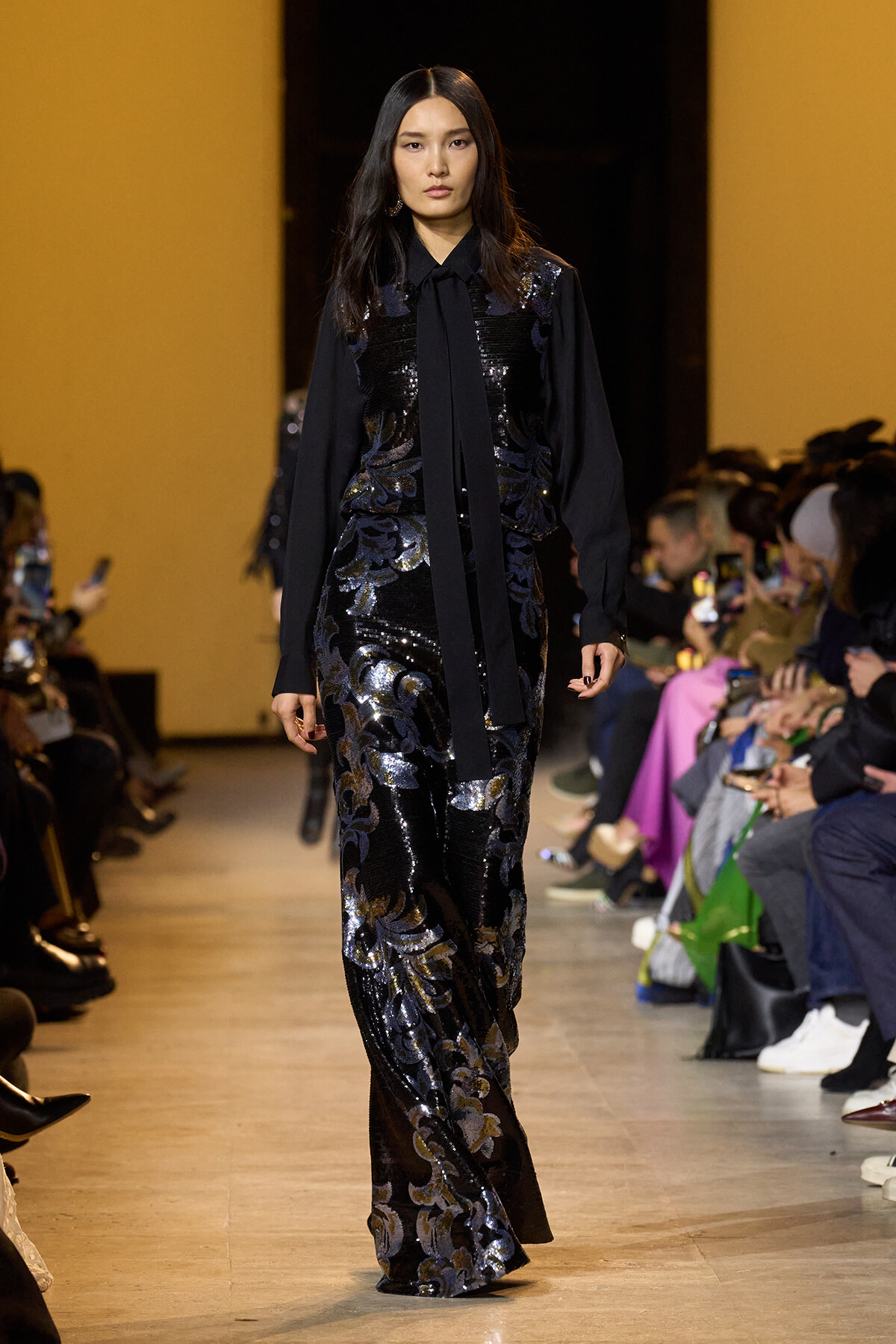 Model on a fashion runway wearing a black sequined dress with a metallic floral pattern and a long black tie, audience on both sides.