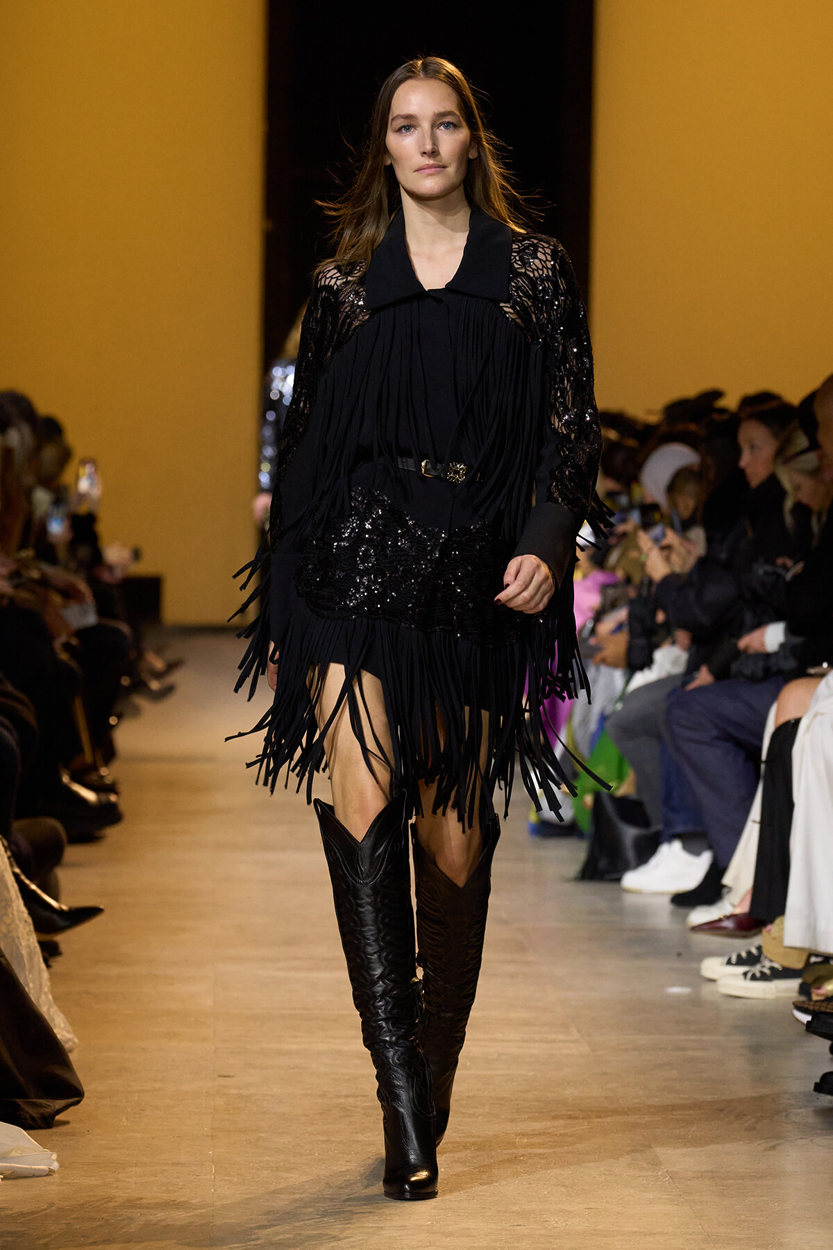 Model walking a fashion runway in a black fringe dress with lace sleeves and knee-high boots, audience on both sides.