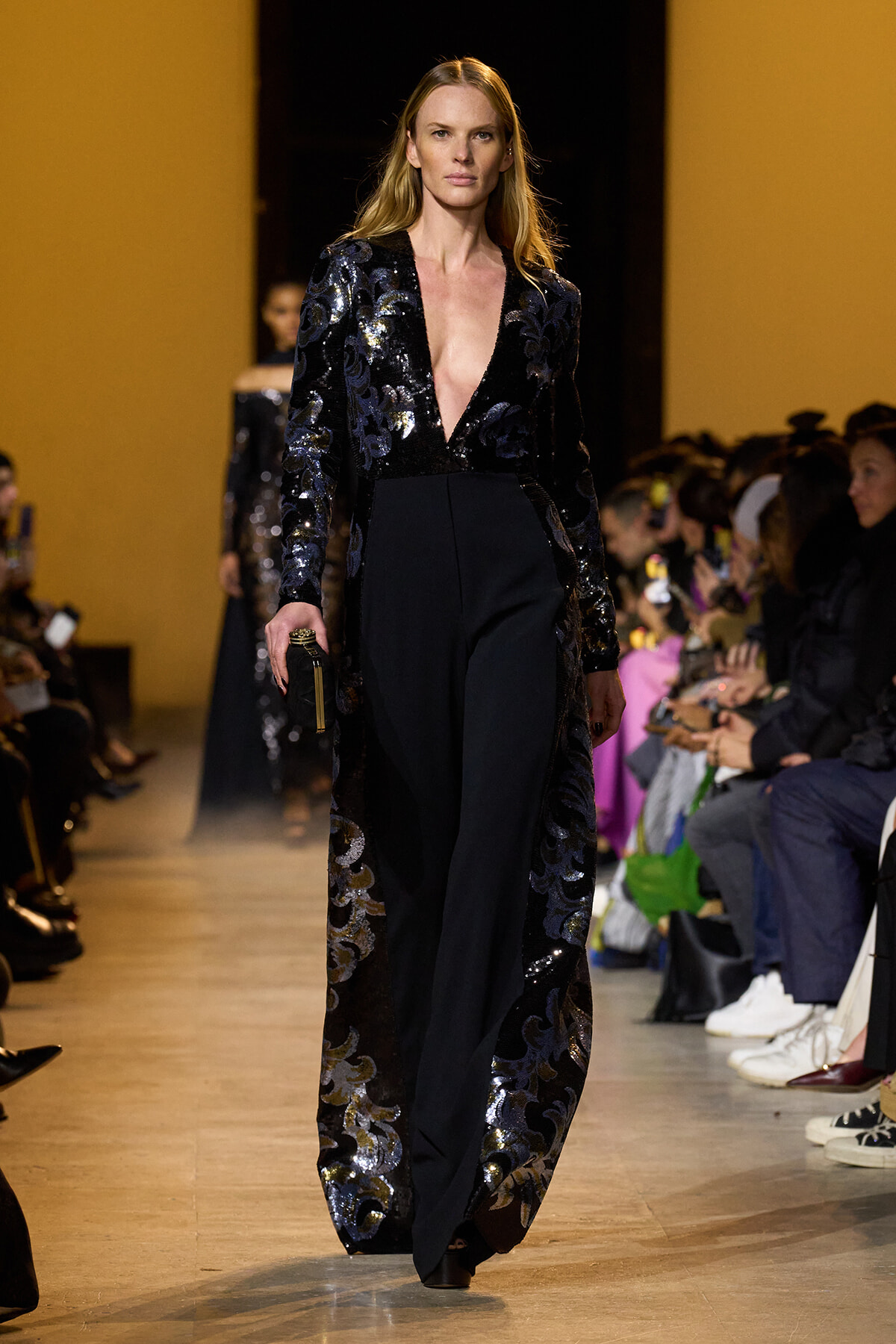 Model walks the runway in a black sequined blazer with a deep V-neck and matching wide-leg pants, holding a small clutch as spectators watch.