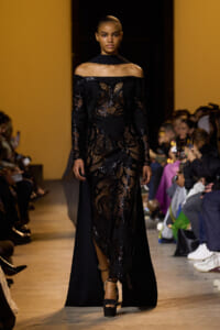 Model walking the runway in a black off-the-shoulder sequin-lace gown with sheer panels and a long cape-like train.