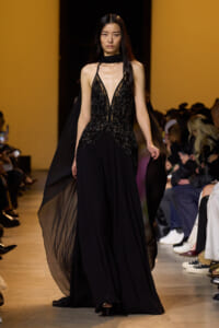 Model walks the runway in a black embellished evening gown with sheer panels and flowing cape, audience on both sides sugaring the spotlight.
