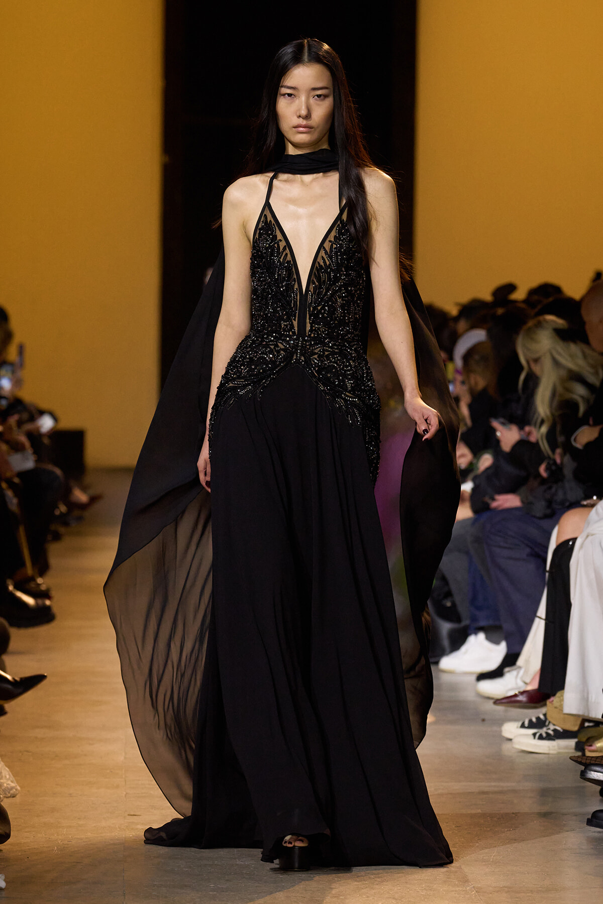 Model walks the runway in a black embellished evening gown with sheer panels and flowing cape, audience on both sides sugaring the spotlight.