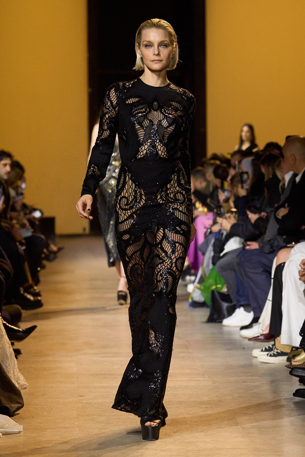 Model walks the runway in a long black lace gown with intricate sheer panels, audience on both sides watching.
