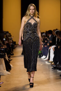 Model on a runway wearing a black, beaded halter gown with intricate geometric embroidery, walking toward the camera.
