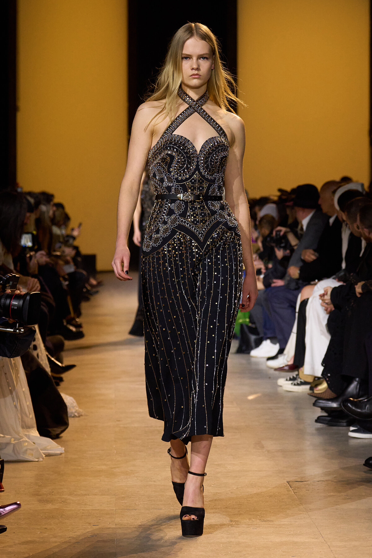 Model on a runway wearing a black, beaded halter gown with intricate geometric embroidery, walking toward the camera.