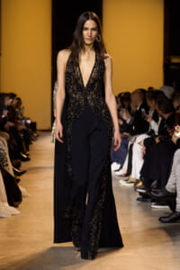 Model walks the runway in a black sequined gown with a deep V-neck and ornate beadwork, audience in the background.