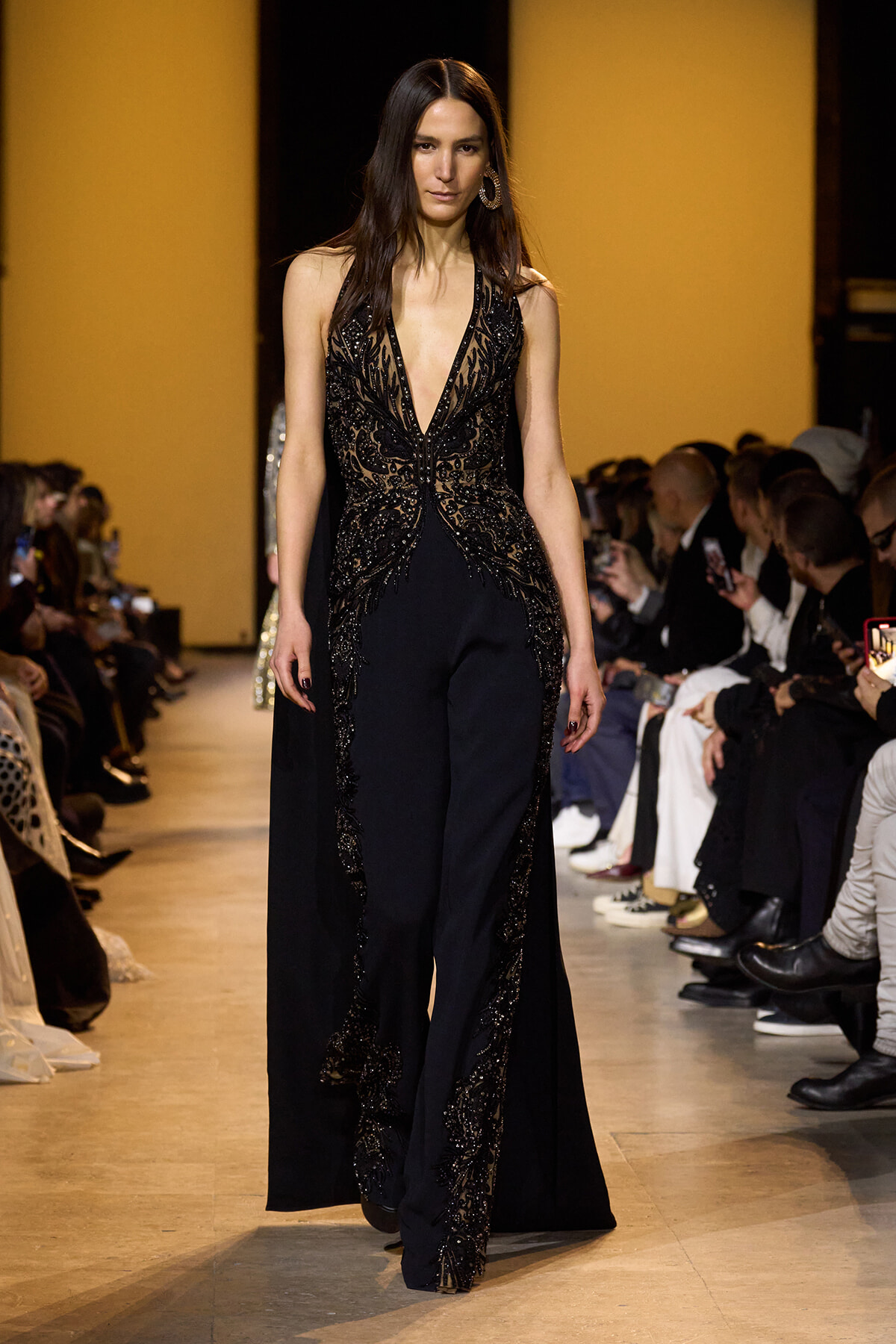 Model walks the runway in a black sequined gown with a deep V-neck and ornate beadwork, audience in the background.