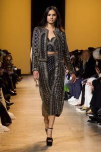 Model walks a fashion runway in a black, gold-embellished crop top, matching jacket and wide-leg pants, with black platform heels.