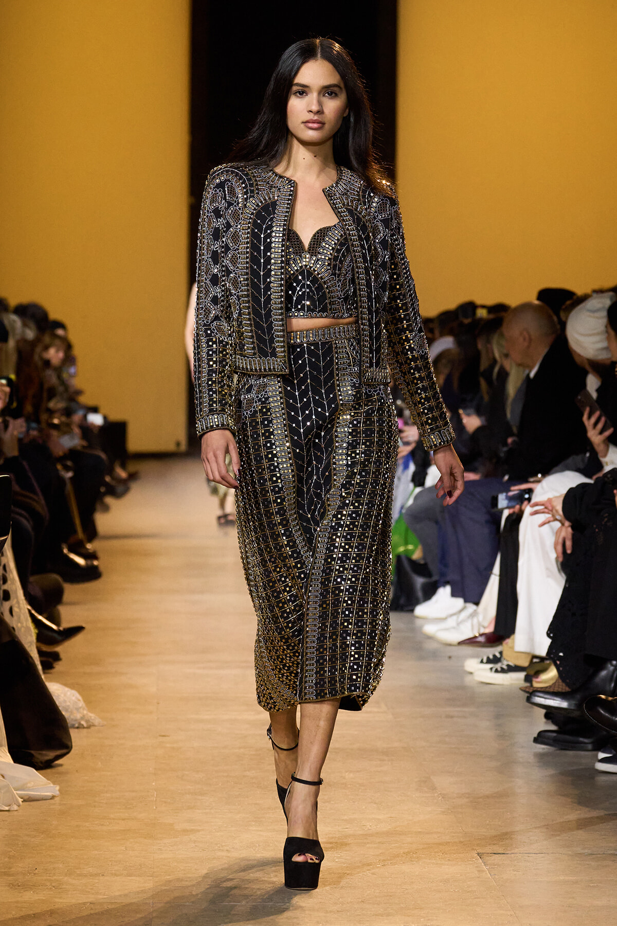 Model walks a fashion runway in a black, gold-embellished crop top, matching jacket and wide-leg pants, with black platform heels.