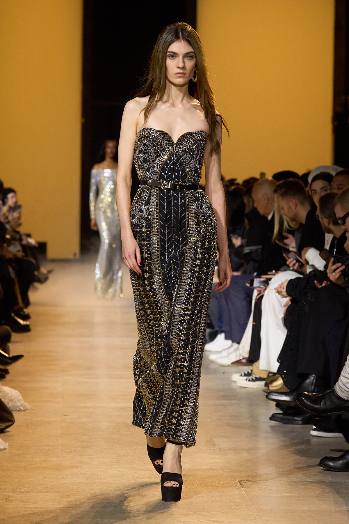 Model walks the runway in a strapless black and gold beaded gown with a geometric pattern; audience seated along the sides.