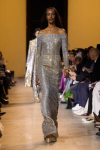 Model walks the runway in a long silver off-the-shoulder sequined gown, audience seated on both sides.