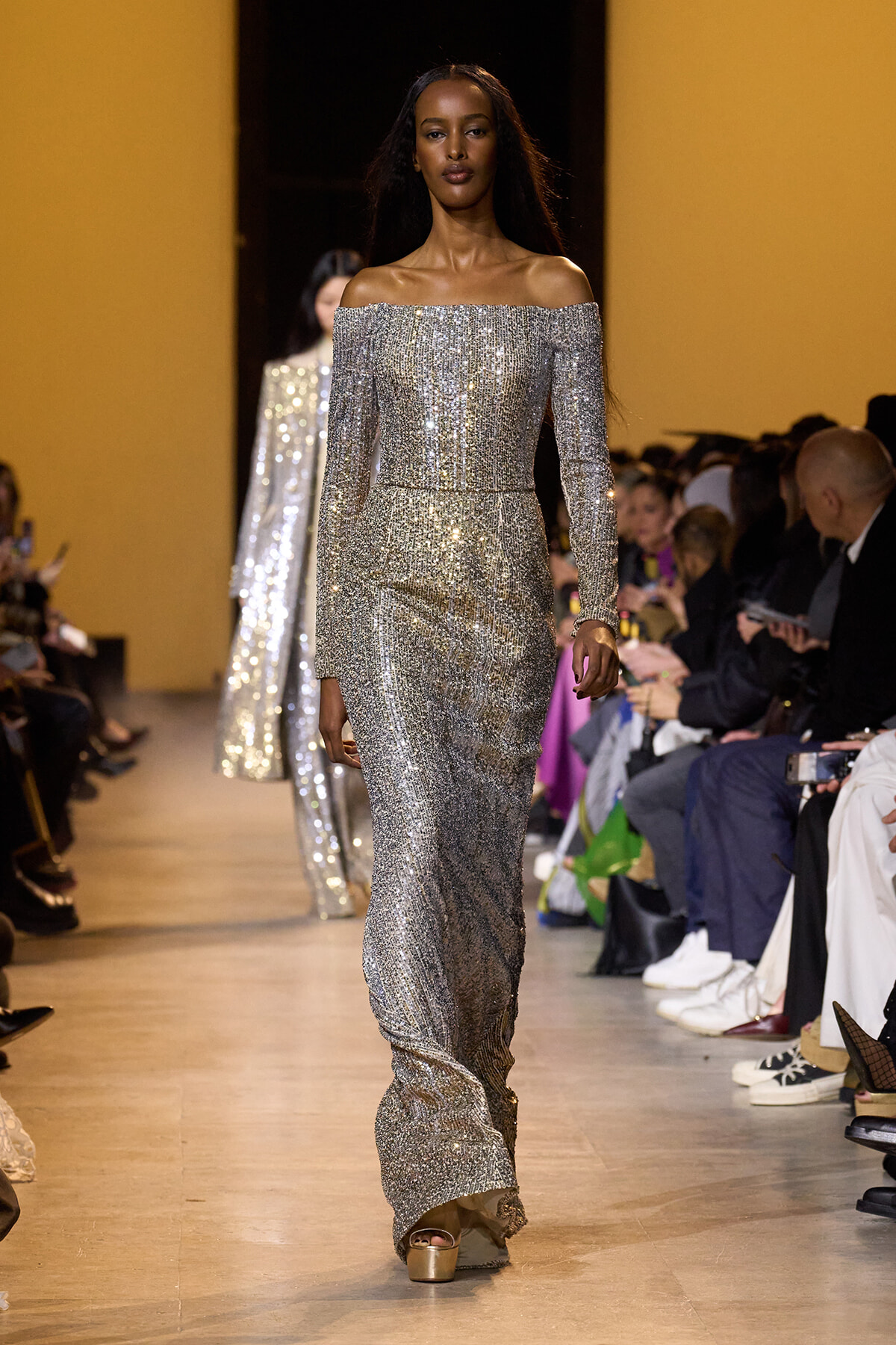 Model walks the runway in a long silver off-the-shoulder sequined gown, audience seated on both sides.