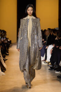 Model walks runway in an oversized silver sequin suit with a pale beige tie and shirt.