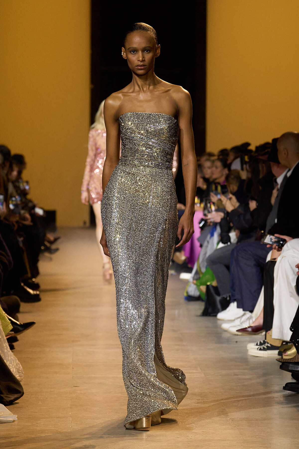 Female model on runway wearing a strapless silver sequined gown with audience watching on both sides.