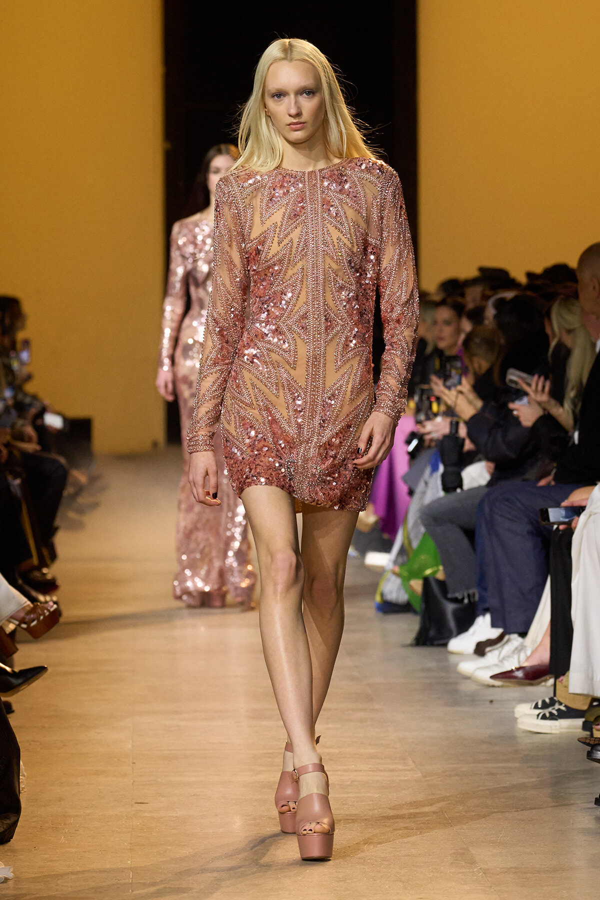 Female model walks the runway in a pink sequin mini dress with long sleeves; audience watches from both sides of the catwalk.