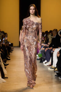 Model walks the runway in a pink sequined off-the-shoulder gown with a large floral embellishment at the shoulder, audience in background.
