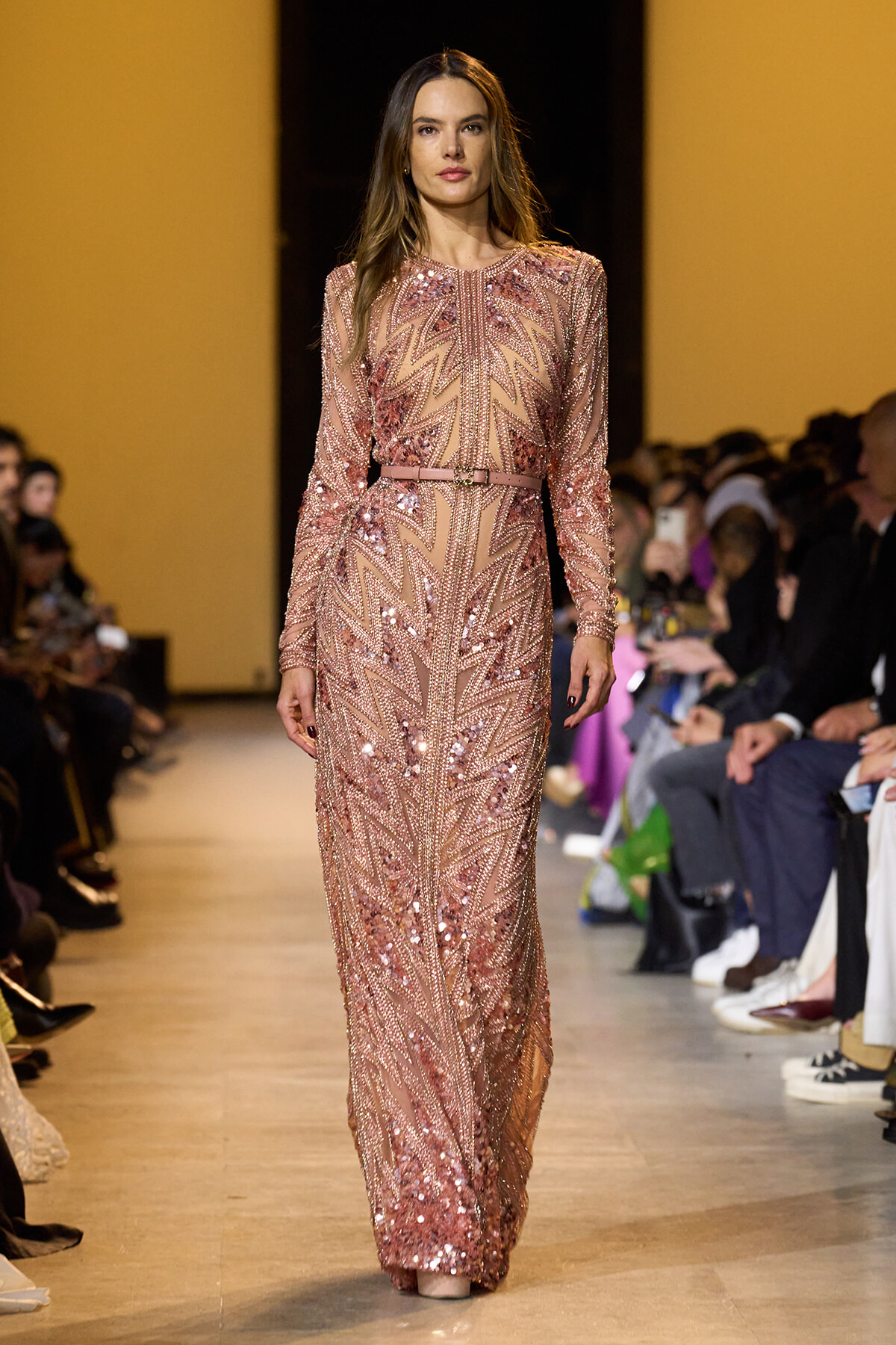 Model walks a fashion runway in a long rose-gold sequin gown with zigzag patterns and a belt at the waist.